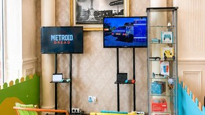 A room with a TV displaying 'METRO DREAD,' a framed cityscape picture, a glass display case with toys, and two chairs on artificial grass.