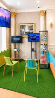 Habitación con una televisión mostrando 'METRO DREAD', una imagen enmarco de un paisaje urbano, una vitrina con juguetes y dos sillas sobre césped artificial.