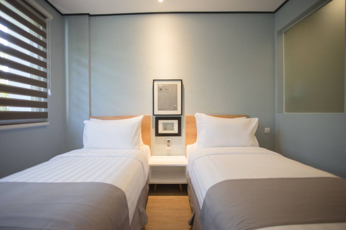 A hotel room with two single beds, a nightstand, a framed picture on the wall, and a window with blinds.