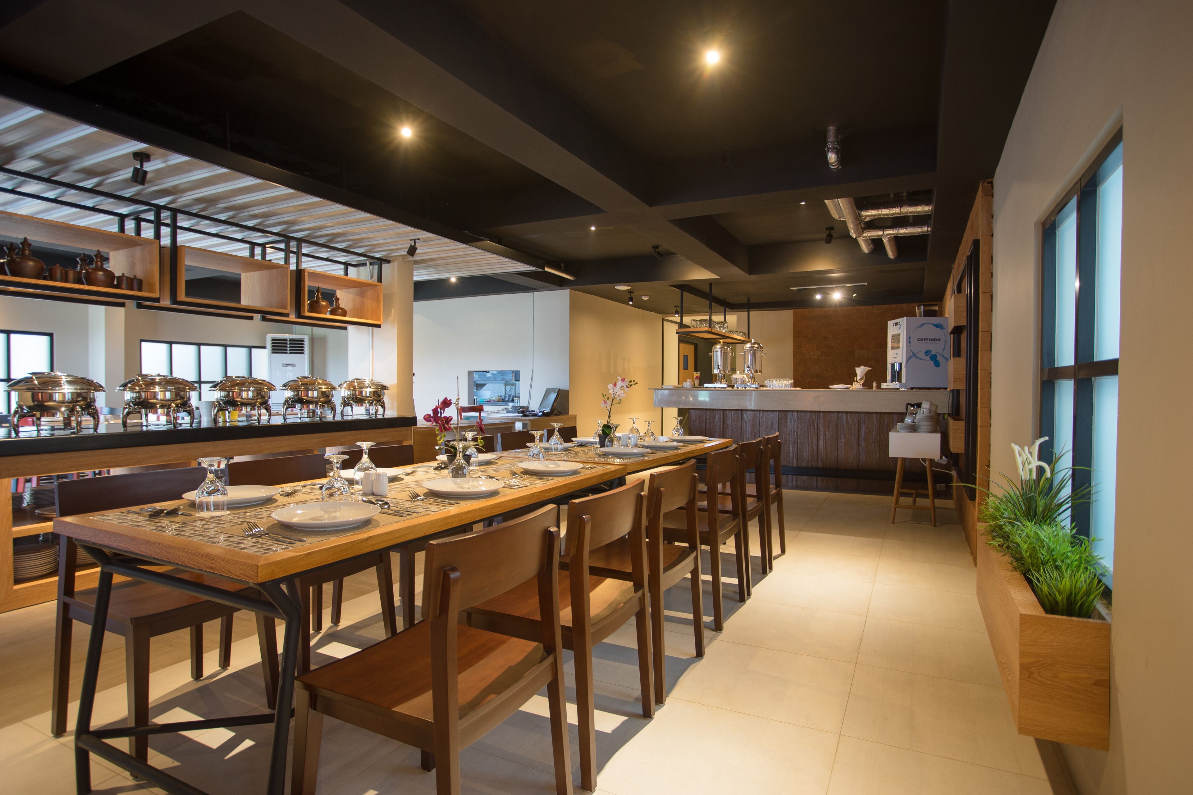 A modern dining area with a long wooden table set for six, surrounded by wooden chairs. There are shelves with kitchenware on the left, a kitchen area in the background, and a potted plant on the right.