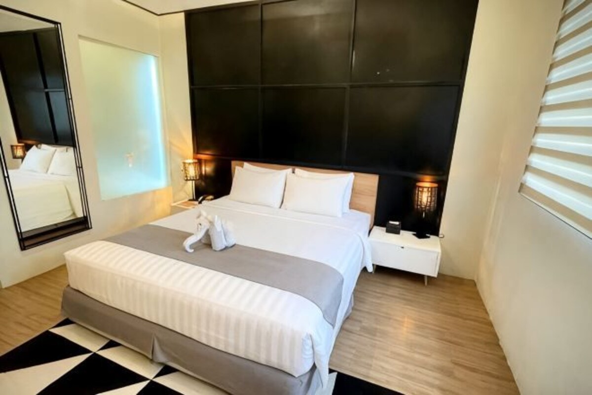 A modern hotel room with a large bed, a nightstand, a mirror, and a wall-mounted headboard.