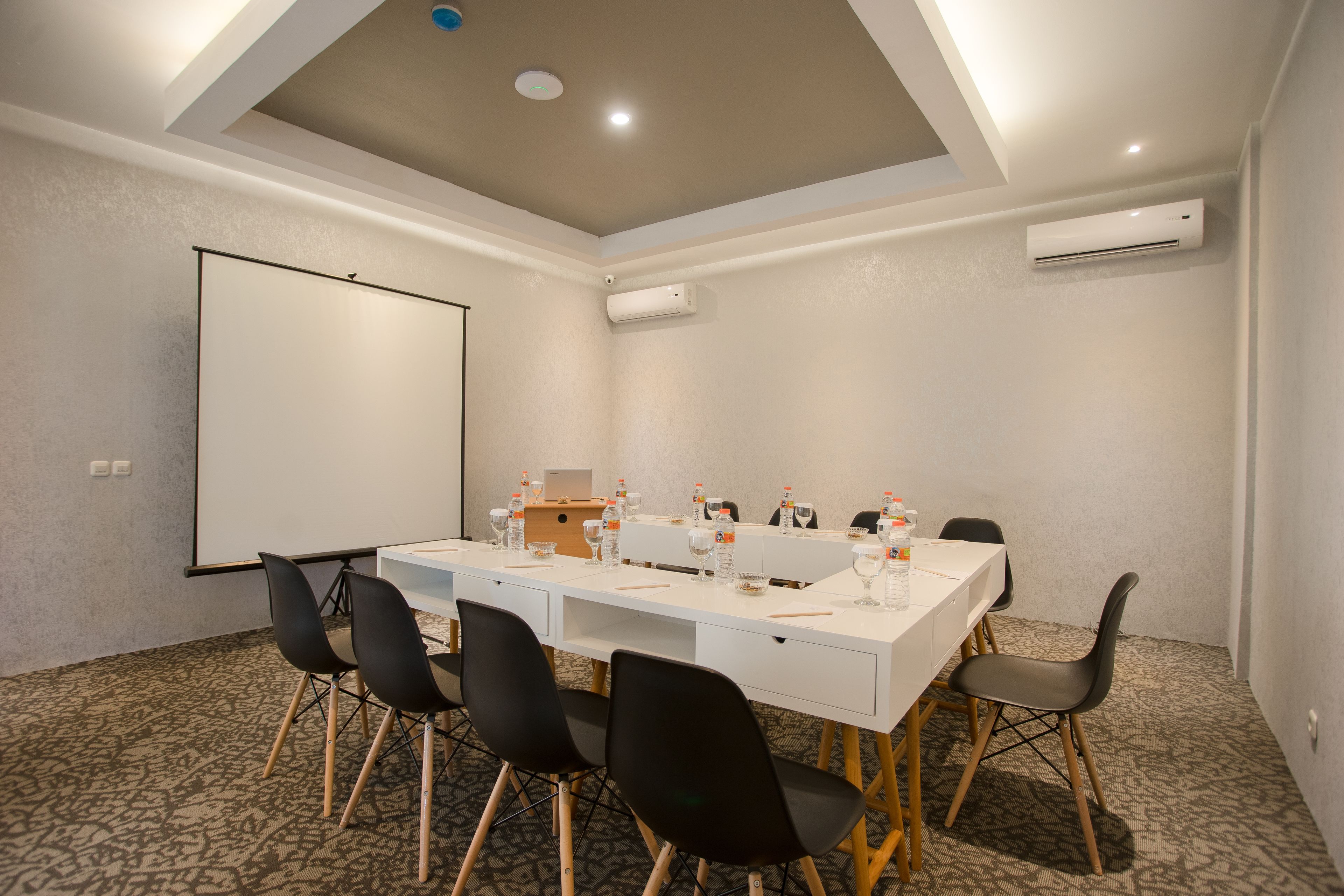 Un salón de conferencias con una mesa blanca, sillas negras, una pantalla de proyector y un equipo de aire acondicionado.