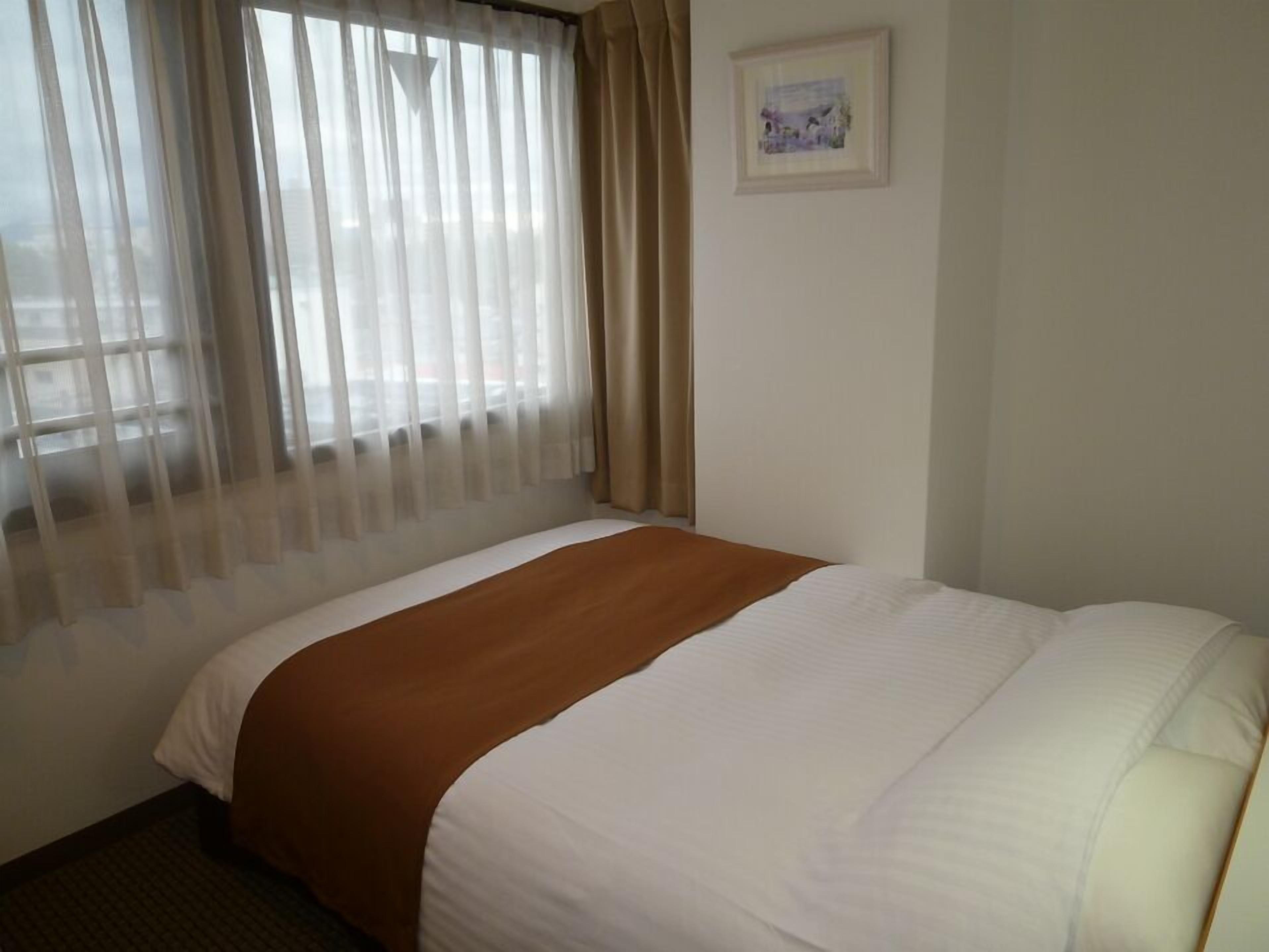 Ein Hotelzimmer mit einem Bett, einem Fenster mit leichtem Vorhang, einem gerahmten Bild an der Wand und einem Teppichboden.