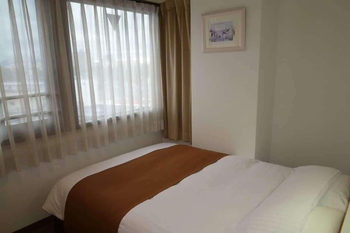 Ein Hotelzimmer mit einem Bett, einem Fenster mit leichtem Vorhang, einem gerahmten Bild an der Wand und einem Teppichboden.