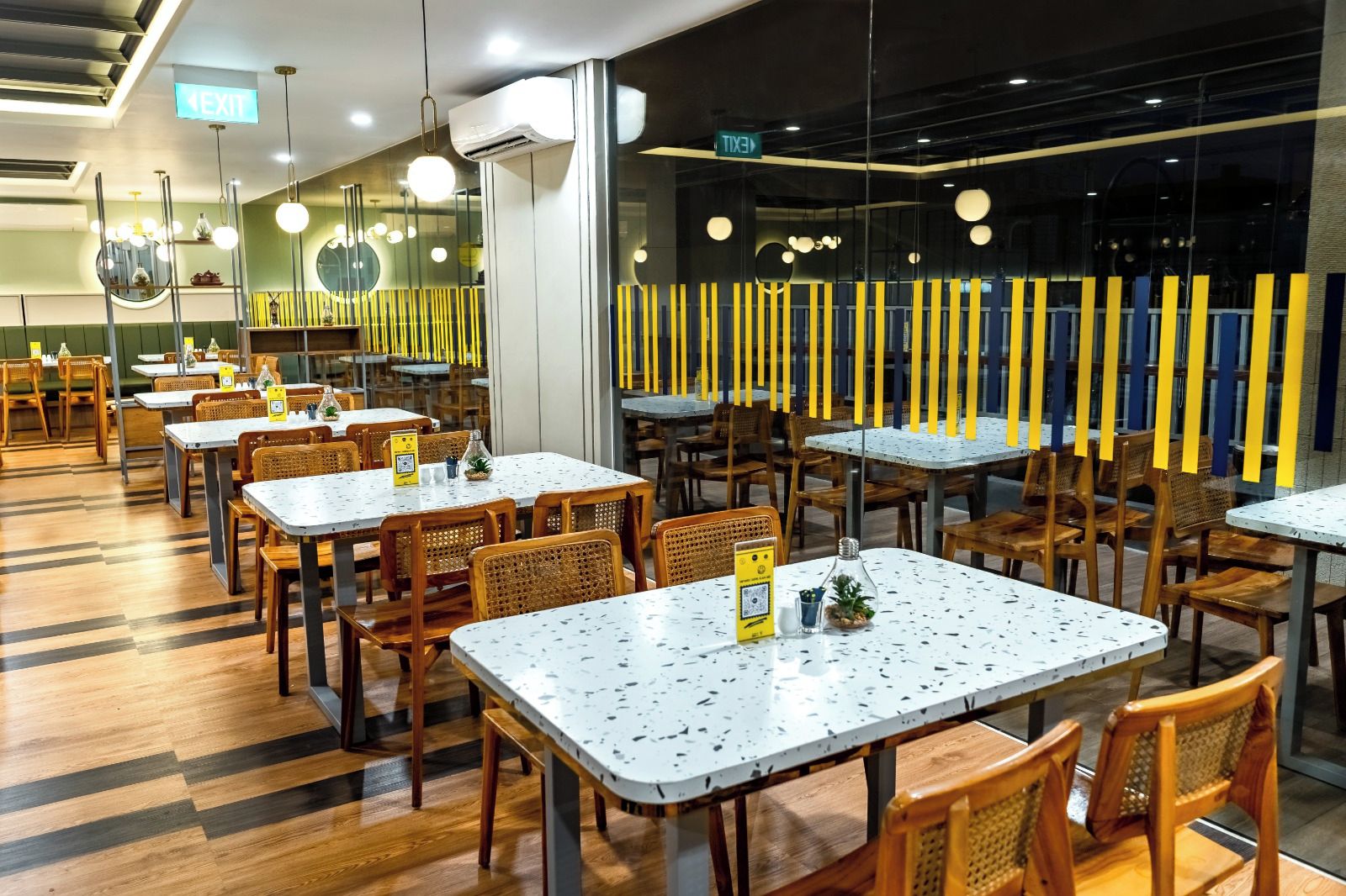 Un interior de restaurante con varias mesas y sillas, un separador de vidrio con elementos verticales amarillos y una señal de salida.