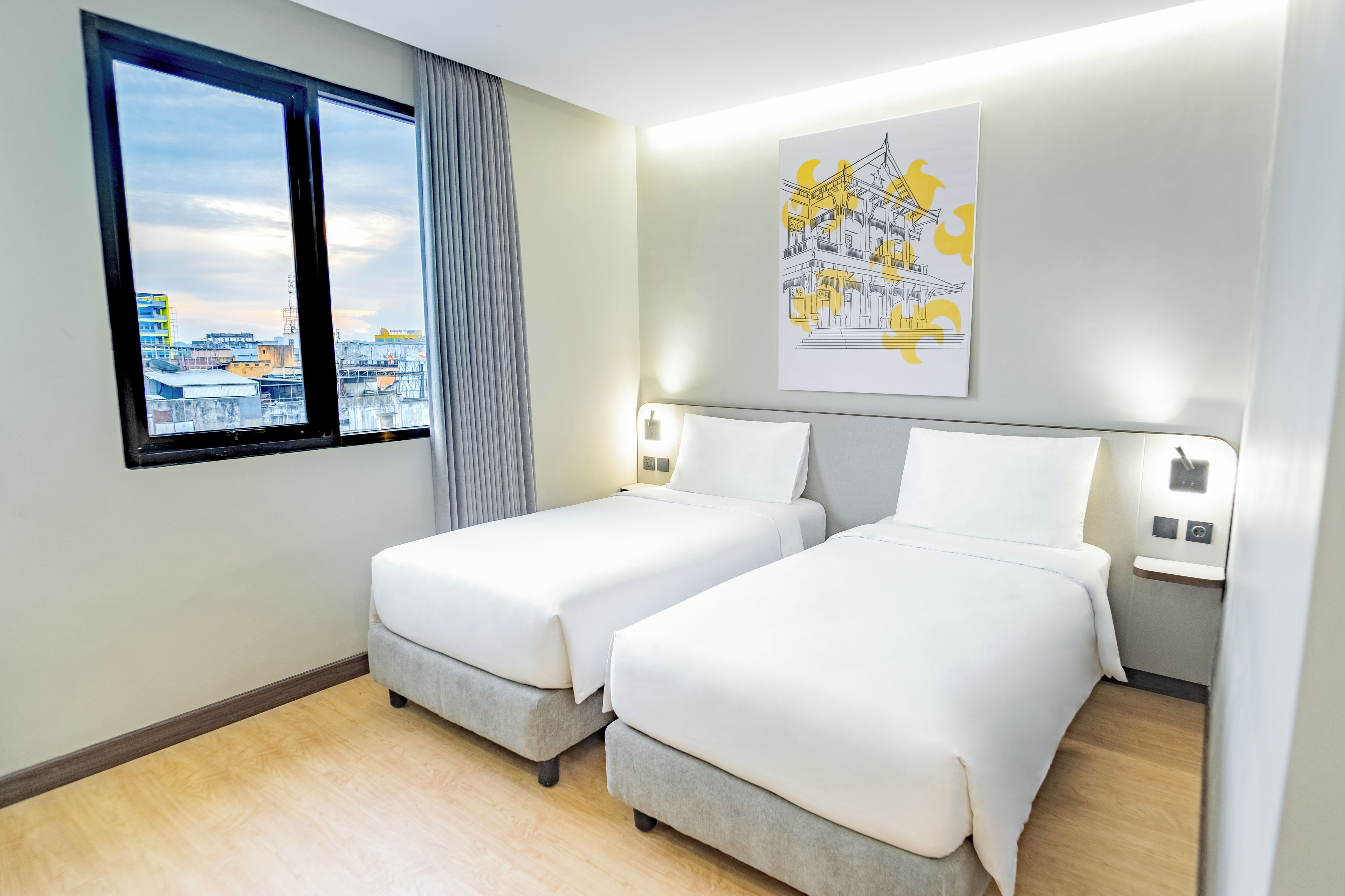 Habitación de hotel con dos camas, ventana con vistas a edificios, una obra de arte colgada en la pared y un interruptor de luz.
