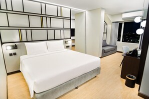 Une chambre d’hôtel moderne dotée d’un grand lit, d’une coiffeuse avec éclairage, d’un coin salon et offrant une vue sur la ville la nuit.