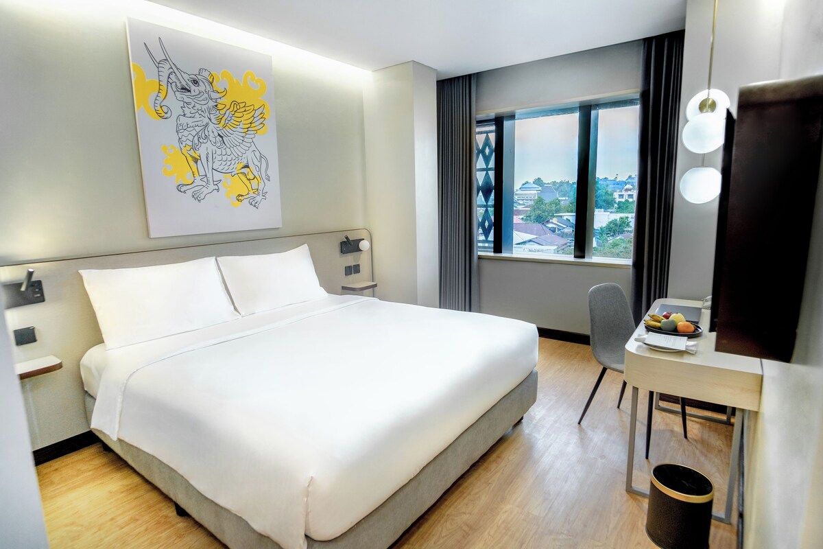 Une chambre d’hôtel avec un grand lit, un bureau avec une chaise, une télévision et une fenêtre donnant sur des immeubles.