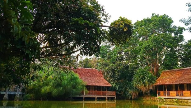 A traditional wooden structure with a tiled roof, surrounded by lush greenery and a calm body of water.