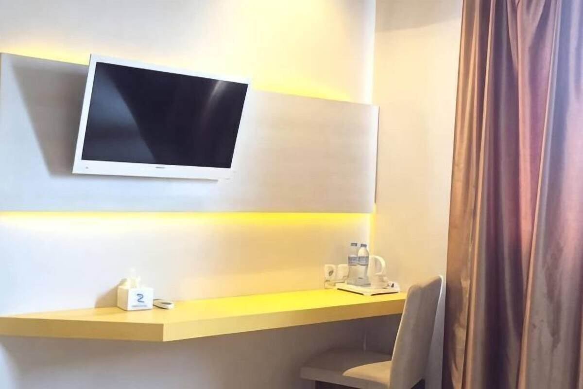 A hotel room with a mounted TV, a desk with a phone and a cup, and a curtain.