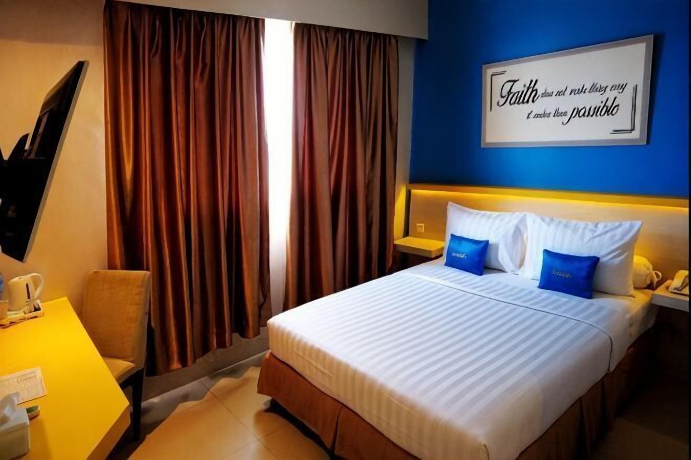 Habitación de hotel con cama, cortinas, escritorio con silla, mesita de noche y una pared con una cita motivacional.