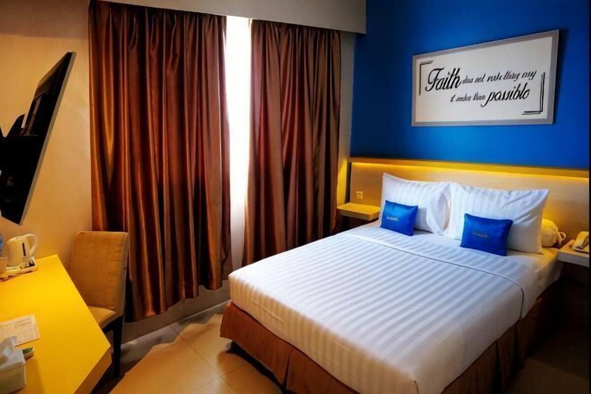 A hotel room with a bed, curtains, a desk with a chair, a nightstand, and a wall with a motivational quote.