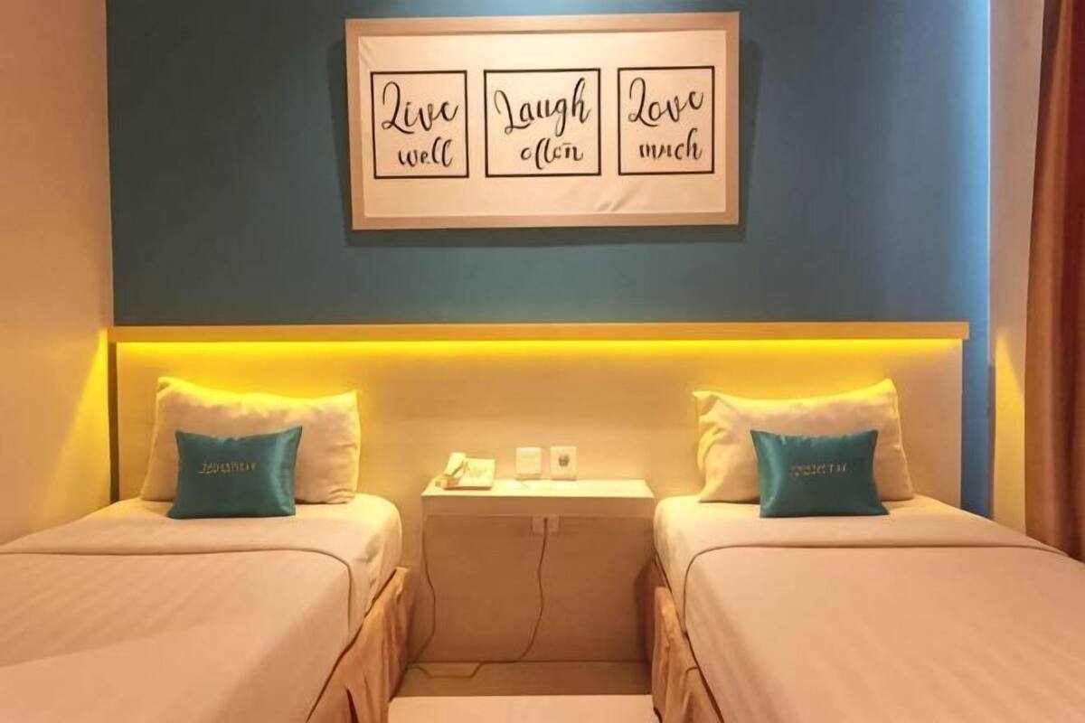 A hotel room with two beds, a bedside table, a wall-mounted sign, and a headboard with integrated lighting.