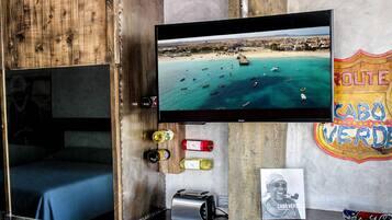 Una habitación con televisión mostrando una escena de playa, pared de madera, cama, una mesita con botellas y un cartel.