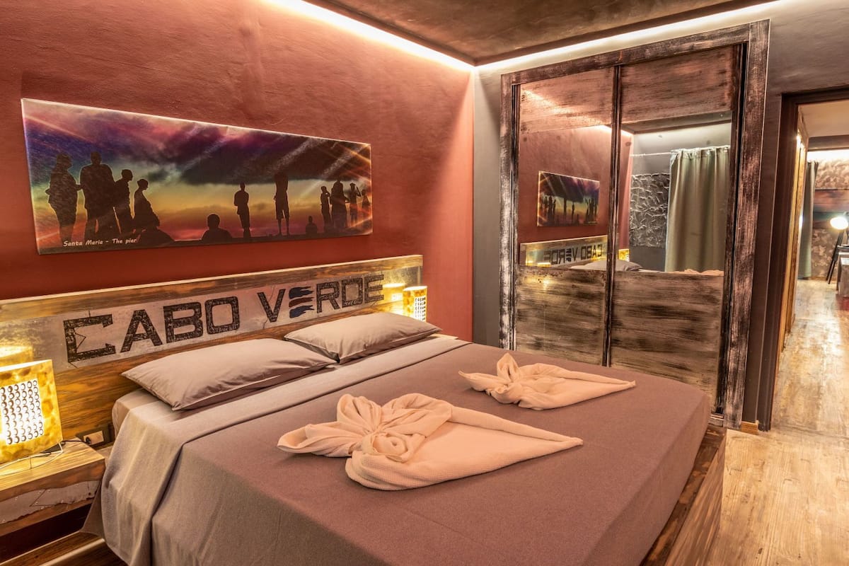 A hotel room with a bed, a headboard with 'CABO VERDE' text, a painting of people on a beach, and a view of the beach through a glass door.