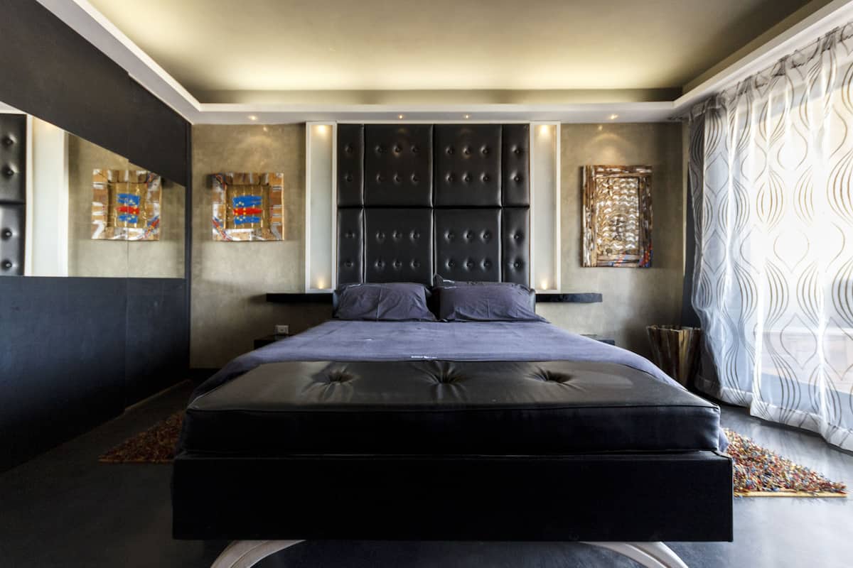 A modern bedroom with a large bed, built-in headboard, and a view of the city through a window.