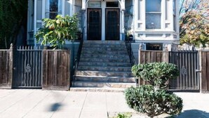 A two-story Victorian-style house with a front porch, a small front yard, and a wooden fence.