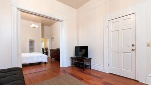 A room with a bed, a television on a stand, a door, and a hallway leading to another room.