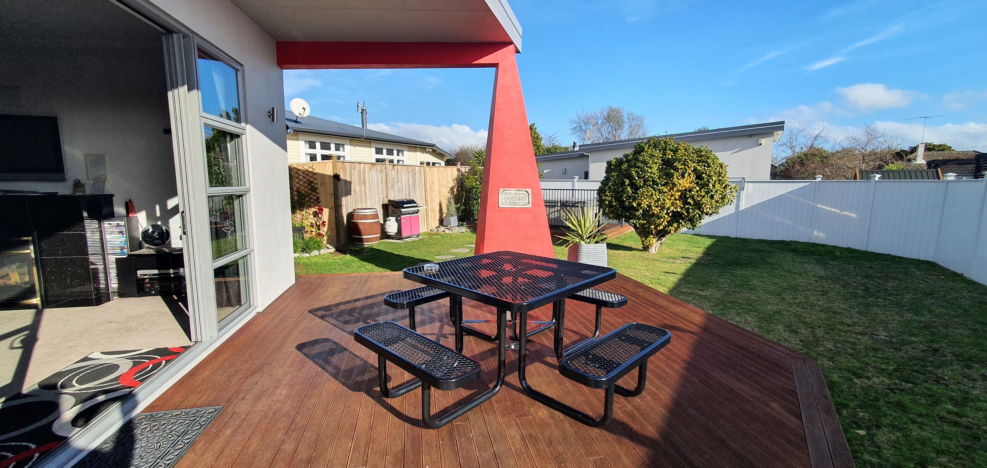 Een terras met een tafel en banken, een houten terras en uitzicht op een achtertuin met een hek en een boom.