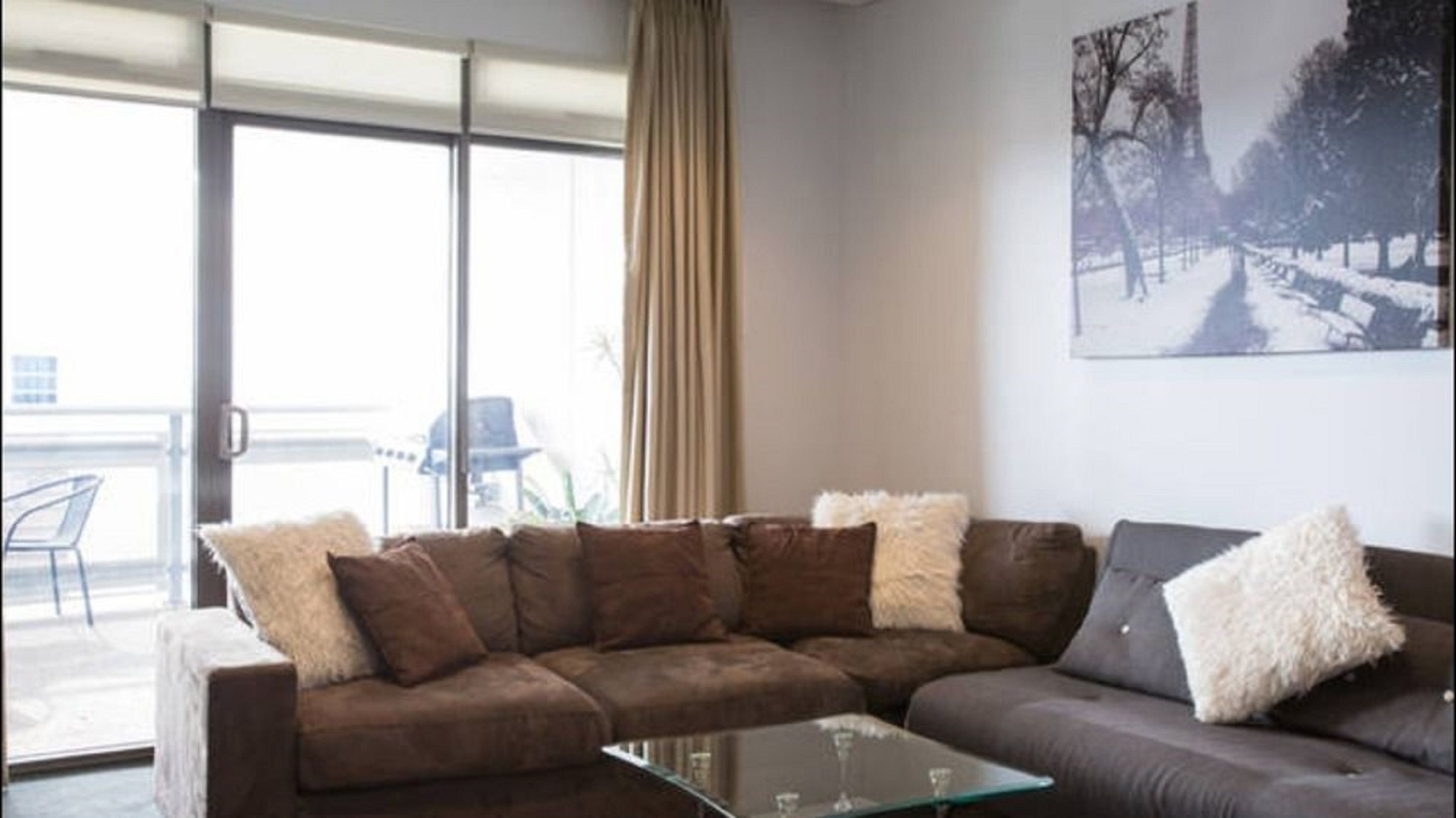 A living room with a brown sofa, a glass coffee table, and a grey sofa. There is a large window with curtains, and a painting of trees on the wall.