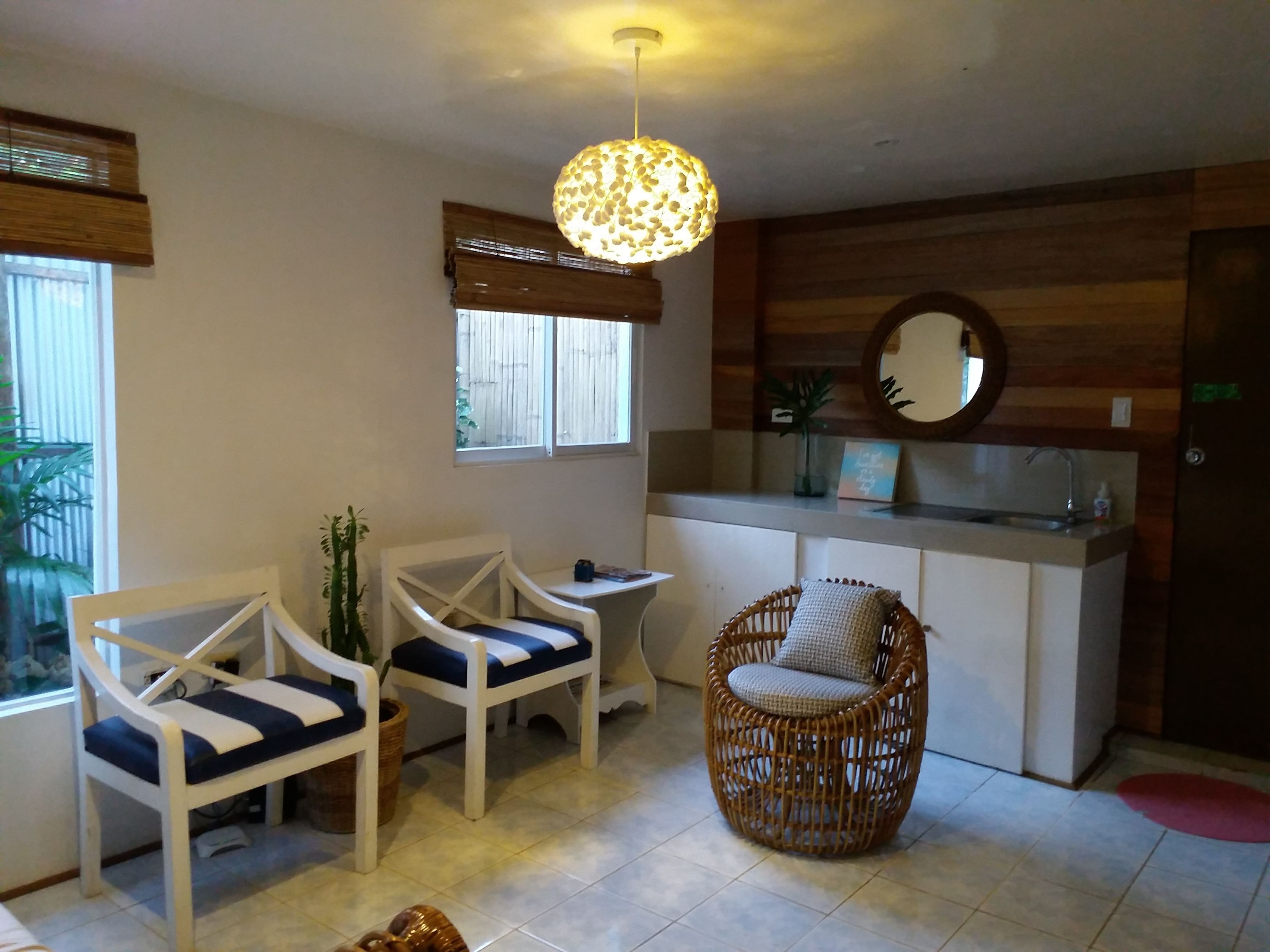 Una habitación con dos sillas blancas con cojines azules, una silla de mimbre, una mesita, una lámpara colgante, una ventana con persianas, un espejo y un lavamanos con una toalla.