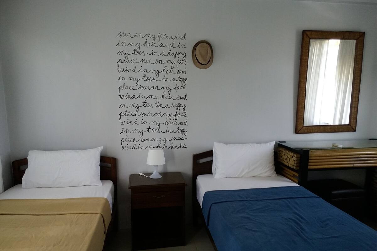 Habitación de hotel con dos camas, una mesita de noche con lámpara, un espejo y una pared con un texto decorativo.