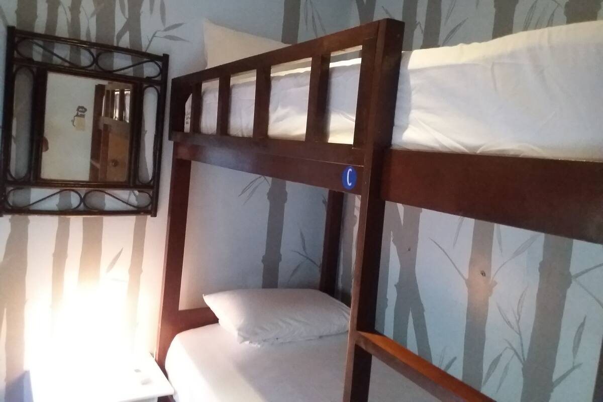 Una cama litera con estructura de madera, colchón blanco y almohada. Junto a la cama hay una mesita con una lámpara.