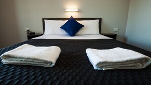 Una cama bien hecha con un edredón oscuro, dos toallas blancas, un almohadón blanco y un almohadón azul decorativo.