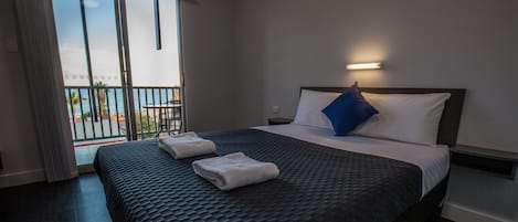 A hotel room with a large bed, a balcony with a view of the sea, and a nightstand with a lamp.