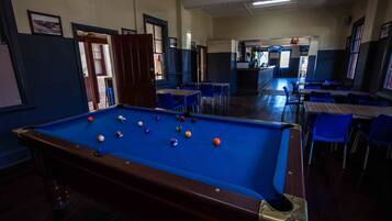 Una mesa de billar con bolas, rodeada de sillas y mesas azules, en una sala con puertas y una barra al fondo.