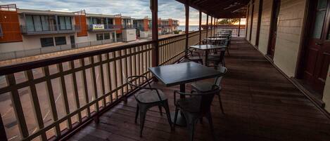 A covered outdoor seating area with tables and chairs, overlooking a street with buildings and a sunset view.