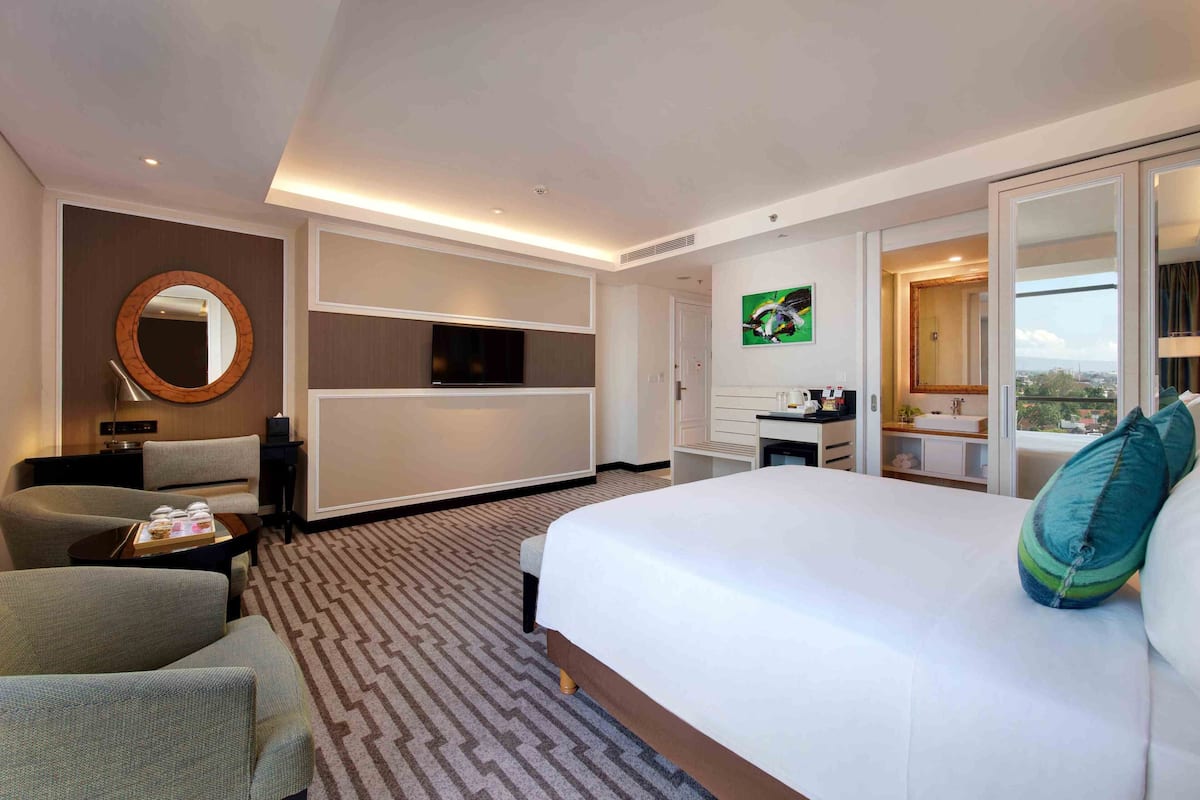 Ein Hotelzimmer mit einem großen Bett, einer Sitzecke mit Sesseln, einem Fernseher, einem Schreibtisch mit Computer und einem Badezimmer, sowie Blick auf die Stadt.