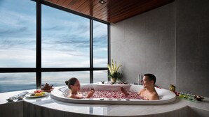 Un baño moderno con una bañera grande llena de pétalos de rosa, un arreglo floral y vistas del cielo a través de amplias ventanas.