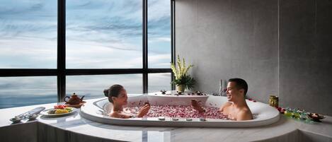 Un baño moderno con una bañera grande llena de pétalos de rosa, un arreglo floral y vistas del cielo a través de amplias ventanas.