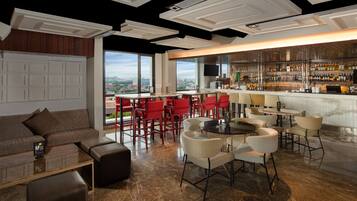 A modern bar area with a view of the city, red bar stools, and a mix of seating options including a couch and round tables.