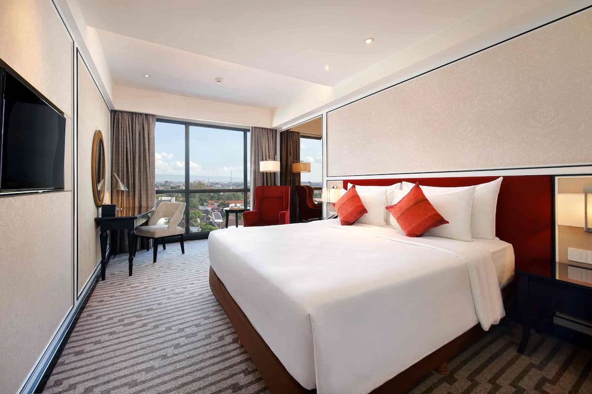 Ein Hotelzimmer mit einem großen Bett, einem Schreibtisch, einem Sessel, einem Fernseher und Blick auf die Stadt.