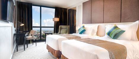 A hotel room with two beds, a desk, a chair, a TV, and a large window with a city view.
