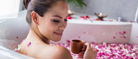 Una persona en una bañera llena de pétalos de rosa, sosteniendo una taza.