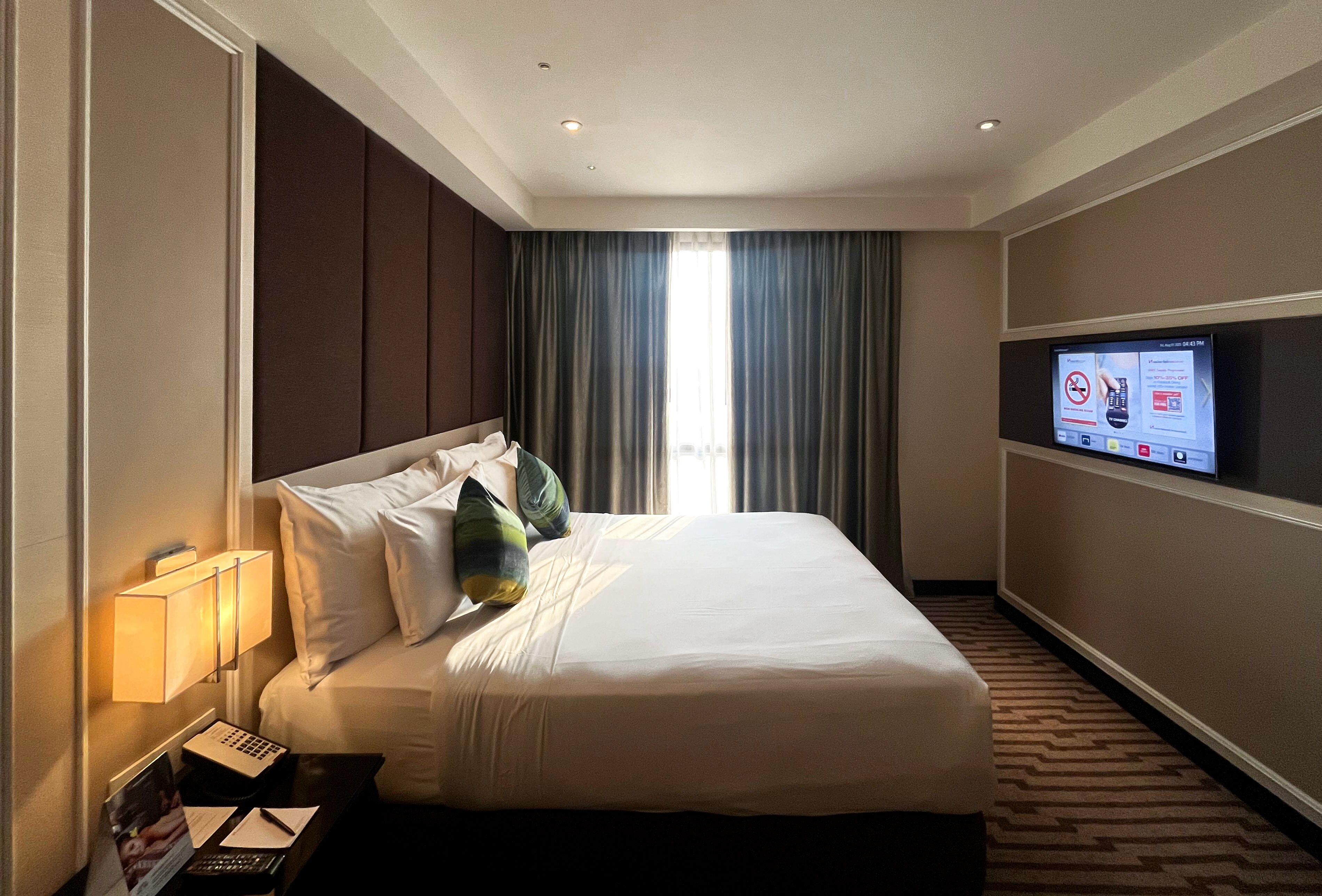 A hotel room with a bed, a TV mounted on the wall, a nightstand with a lamp, and a window with curtains.