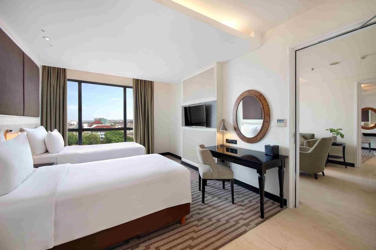 Ein Hotelzimmer mit zwei Betten, einem Schreibtisch, einem Fernseher und einem großen Fenster mit Blick auf die Stadt.