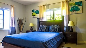 A bedroom with a large bed, two bedside tables with lamps, a window with curtains, and artwork on the walls. - Skyview Manor Guest Apartments (Kingston)