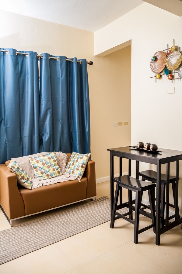 Una habitación con un sofá marrón, una mesita con taburetes, una cortina azul y un adorno en la pared.