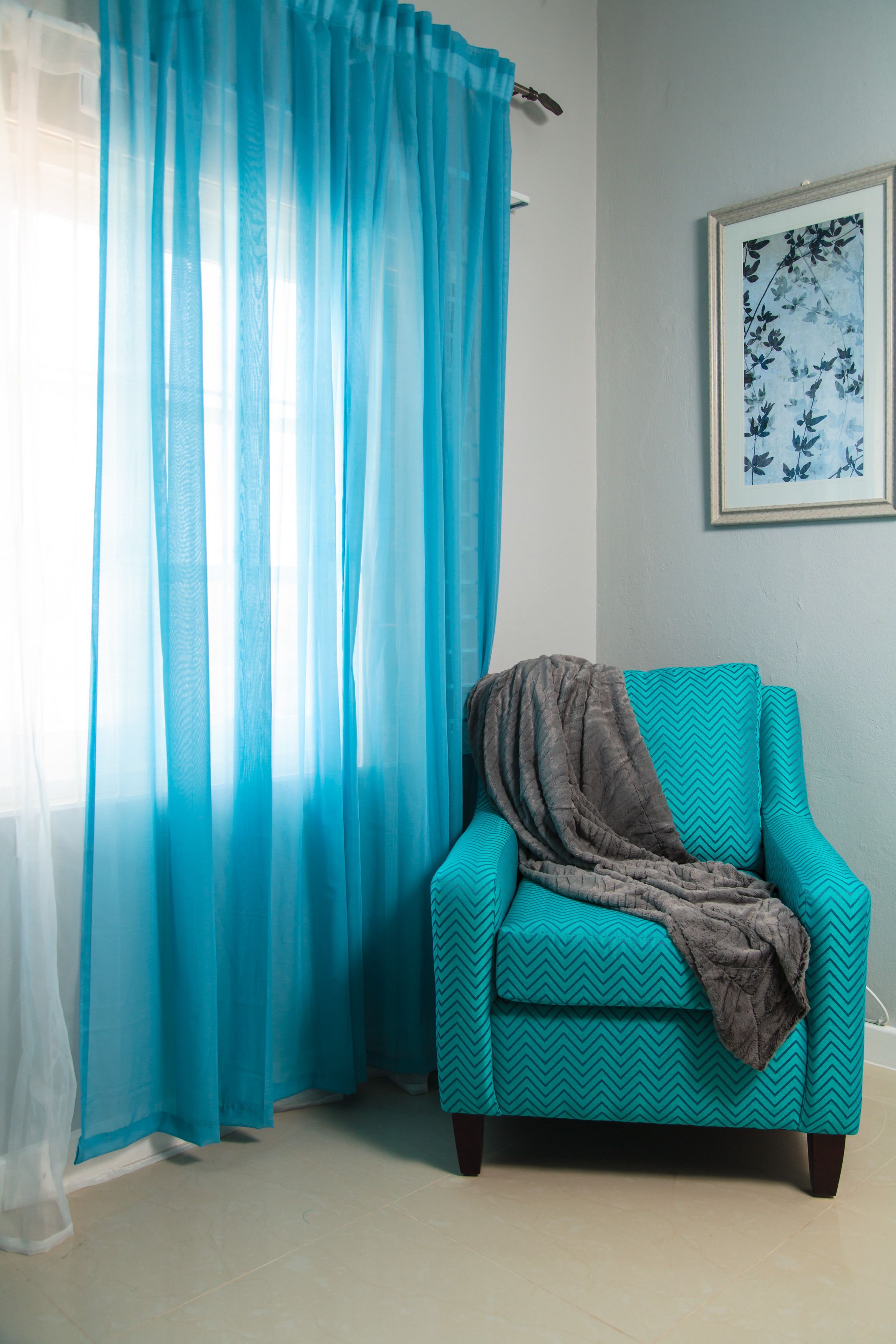 Un fauteuil turquoise agrémenté d’un plaid à motifs, un rideau bleu transparent et une œuvre d’art florale encadrée accrochée au mur.