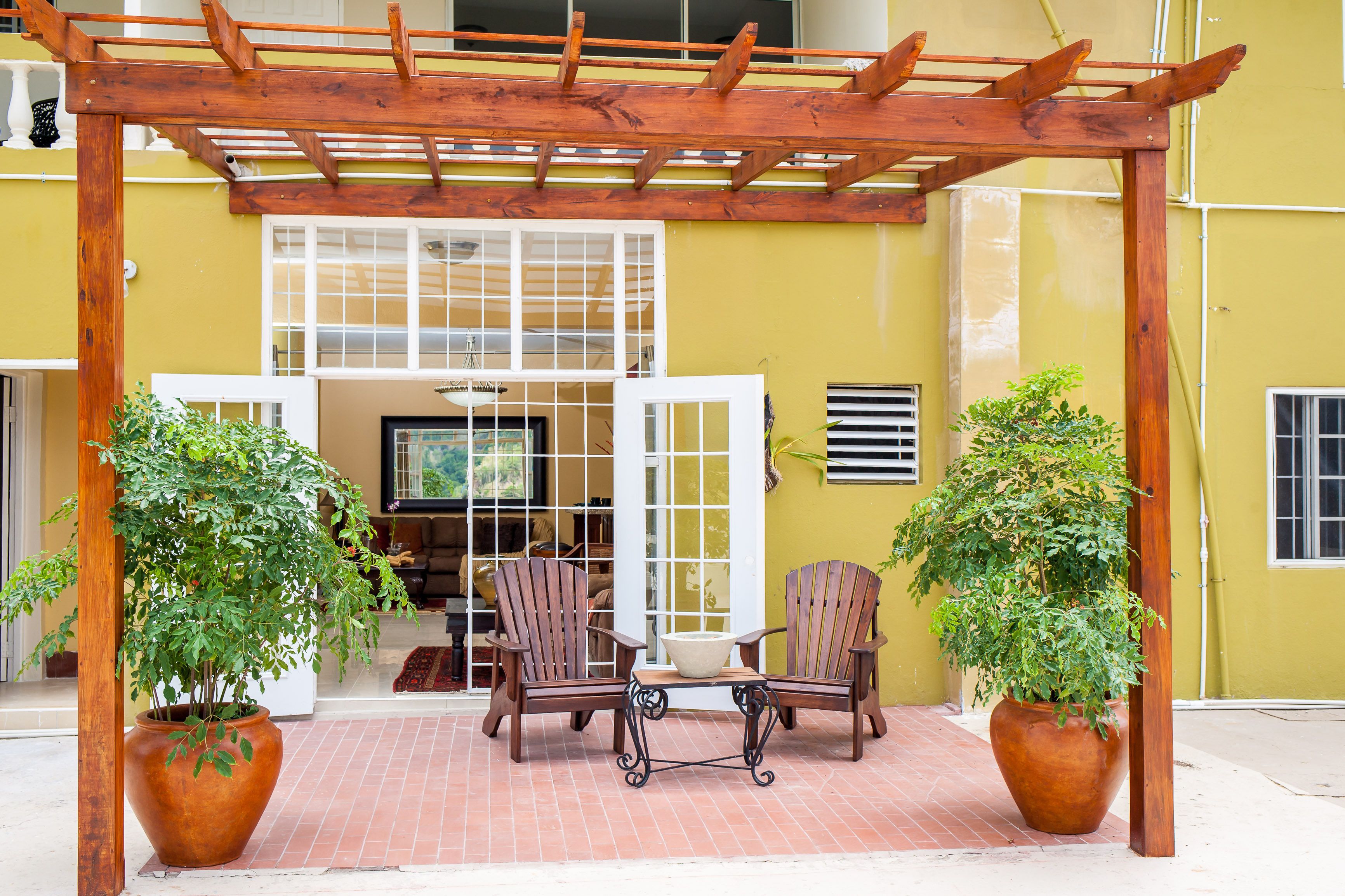Une pergola en bois, agrémentée de deux chaises et d’une table, entourée de plantes en pot, se trouve devant un bâtiment jaune doté d’une grande fenêtre et d’un téléviseur.