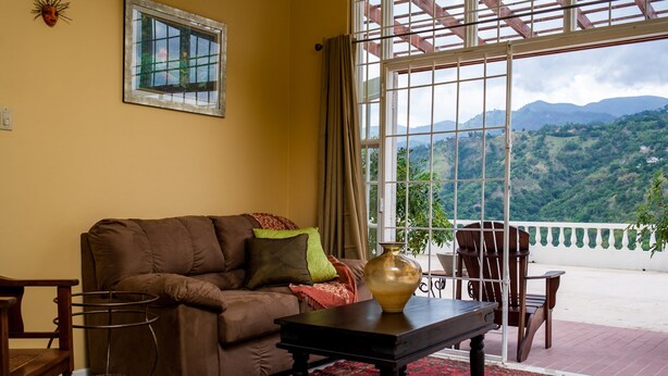 A living room with a brown sofa, a black coffee table, and a large window offering a view of green hills. - Skyview Manor Guest Apartments (Kingston)