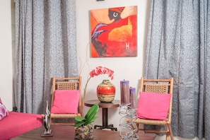 Une pièce avec une table en bois, deux chaises en osier dotées de coussins roses, un vase avec des fleurs rouges et un tableau accroché au mur.