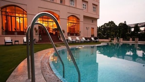 Uma piscina com guarda-corpo metálico curvo, um gramado bem cuidado e um edifício com grandes janelas ao fundo.