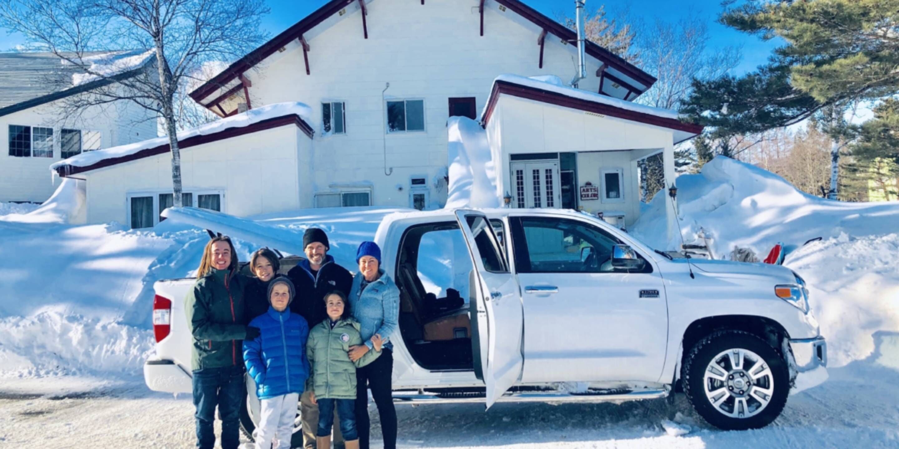 Eine Gruppe von Personen steht vor einem weißen Gebäude mit einem verschneiten Einfahrt und einem weißen Pickup-Truck mit geöffneter Tür.
