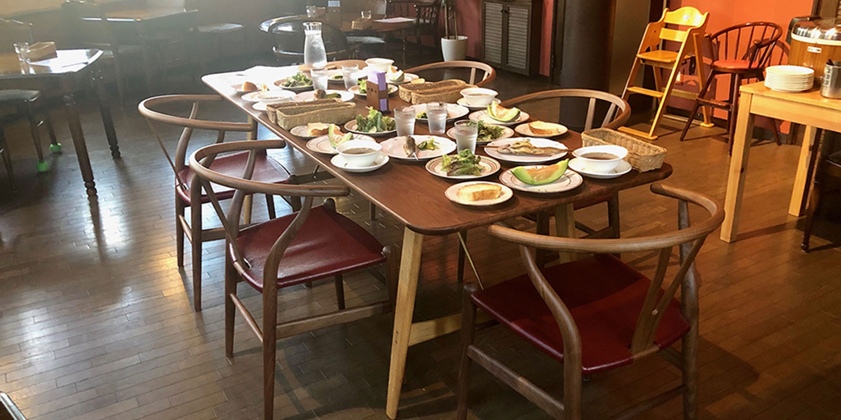 A dining area with wooden tables and chairs, various dishes and drinks on the tables, and a potted plant in the background.