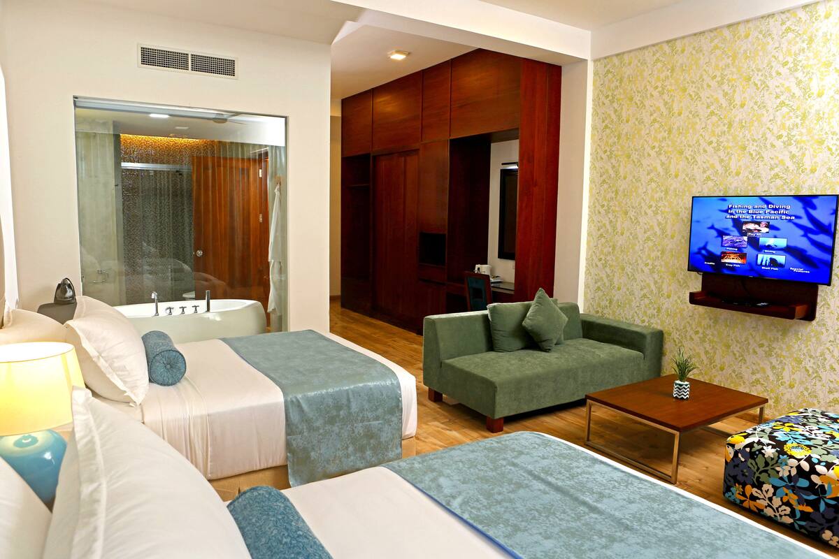 Une chambre d’hôtel avec deux lits, un canapé vert, une petite table et un téléviseur diffusant une émission.